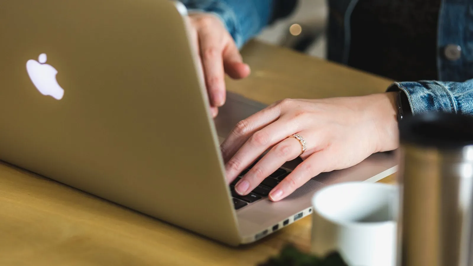 Mãos digitando no teclado de um laptop sobre uma mesa de madeira, ao lado de uma garrafa térmica