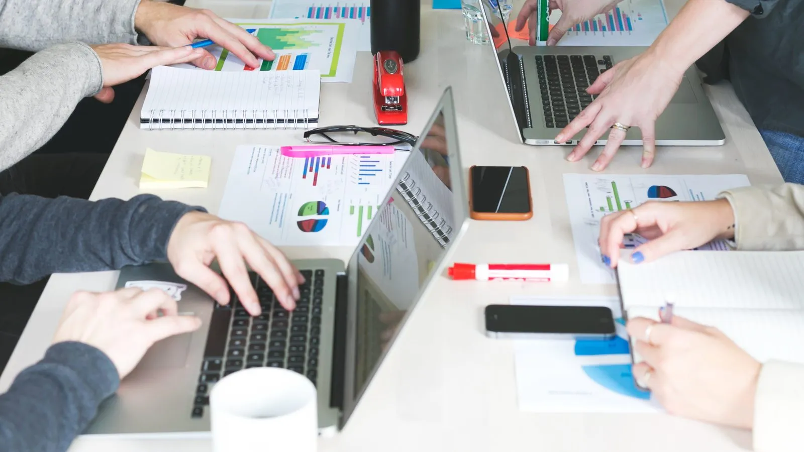 Várias pessoas ao redor de uma mesa de trabalho, com laptops, gráficos impressos, cadernos e canetas marcadoras