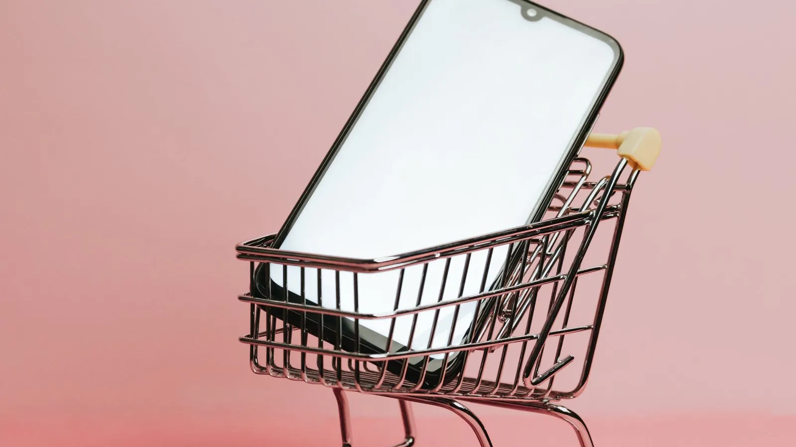 Um celular com a tela em branco dentro de um carrinho de compras em miniatura, sobre fundo rosa
